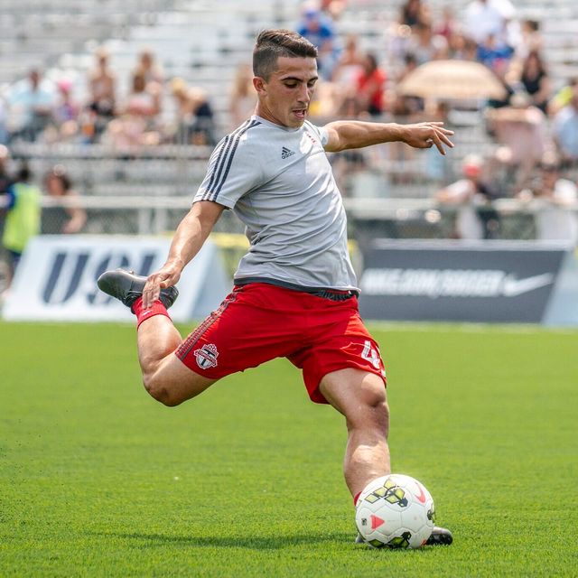 Luca in a grey warm-up top and red shorts striking the ball, a banner behind.